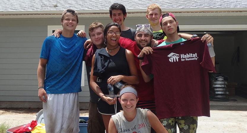 A group of young people pose for a photo. One of them is holding a habitat for humanity shirt.